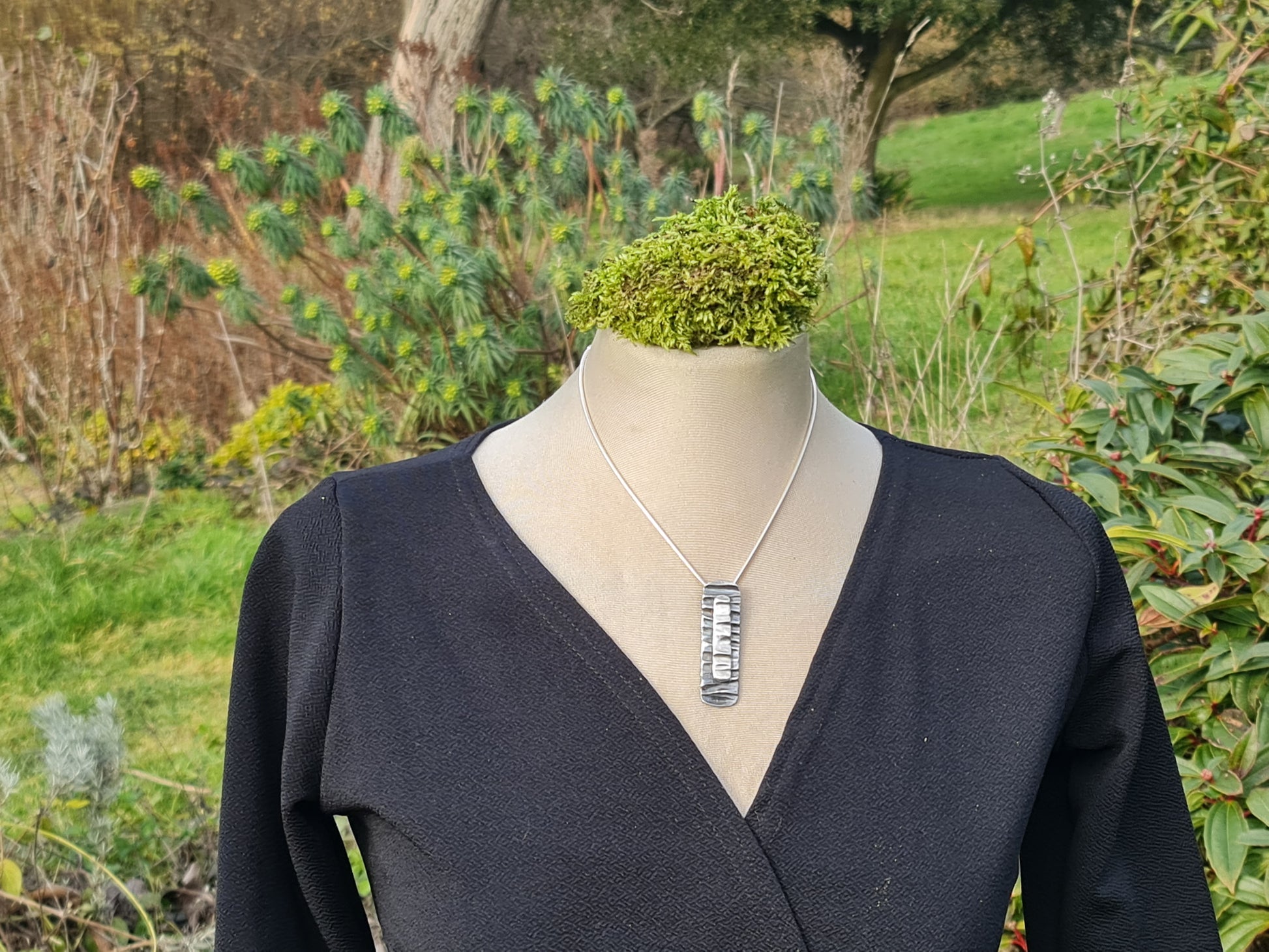 Handmade tree bark textured sterling silver rectangle with curved ends with a smaller, same shaped sterling silver shape on top.  The underneath tree bark shape is oxidised and the top one is polished to give a two tone effect.  The pendant is on a snake chain and can be 16, 18 or 20".  The pendant is on a mannequin wearing a black dress and is shown with a garden background.