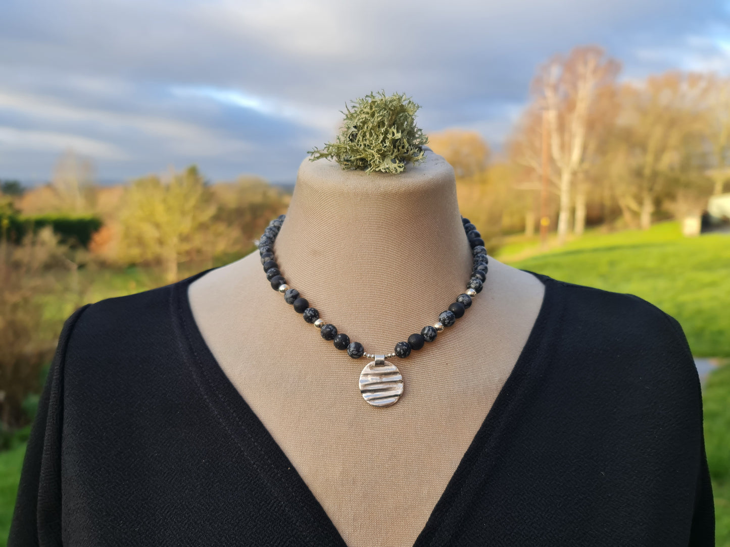 Black snowflake obsidian and textured sterling silver circle necklace with silver beads. Shown on a mannequin with a black dress and the background is a country english garden