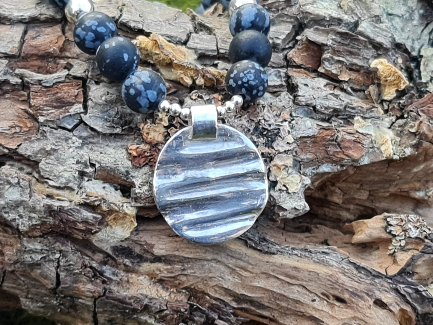 Close up of ripple textured sterling silver circle hanging from small silver beads and the necklace is black snowflake obsidian.  The necklace is shown on a tree log.