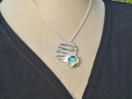 handmade sterling silver rippled pendant with a beautiful blue and green paua shell placed off centre.  The pendant is shown on the top half of a mannequin wearing a black dress