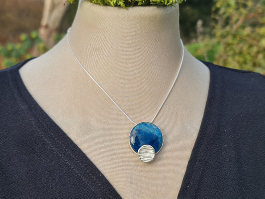 3 cm Blue apatite cabochon with a 1 cm concave textured sterling silver cirlce offset pendant on a snake chain.  The handmade pendant is on a mannequin set in the garden.