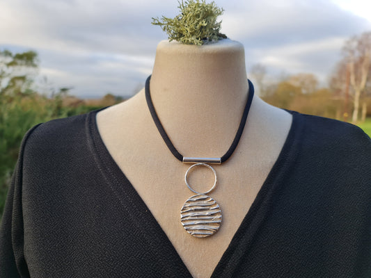 A truly gorgeous sterling silver necklace with a solid tree bark textured circle and a hollow circle on top and a silver tube which the black silk goes through.  The necklace is shown on a mannequin with a countryside background.