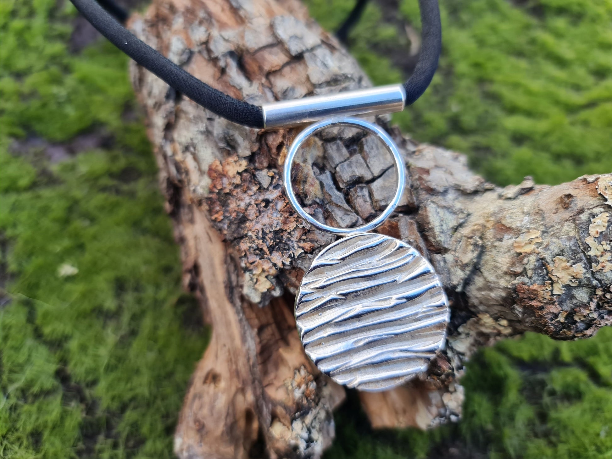 circular sterling silver pendant with a tree bark texture is attached to a smaller hollow circle.  The pendant has a silver tube which the black silk runs through and attaches with a silver clasp.  The pendant is shown on log with moss in the backround.