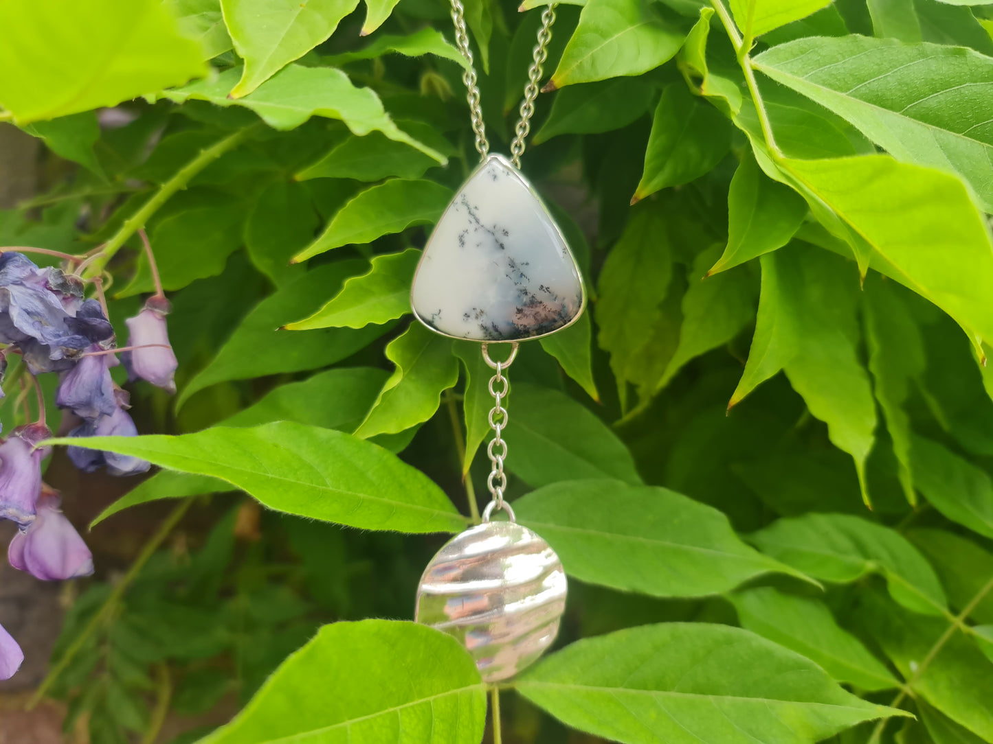 Dendritic opal in a triangular shape with a domed sterling silver disk.  A pendant on a silver chain in front of a wisteria tree