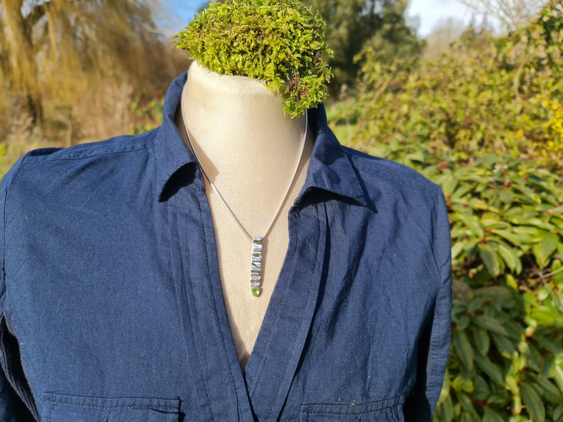 Tree Bark textured sterling silver rectangular pendant with a bright green triangular peridot at the bottom of the pendant.  The pendant is on a mannequin with a blue shirt and set in a garden scene.