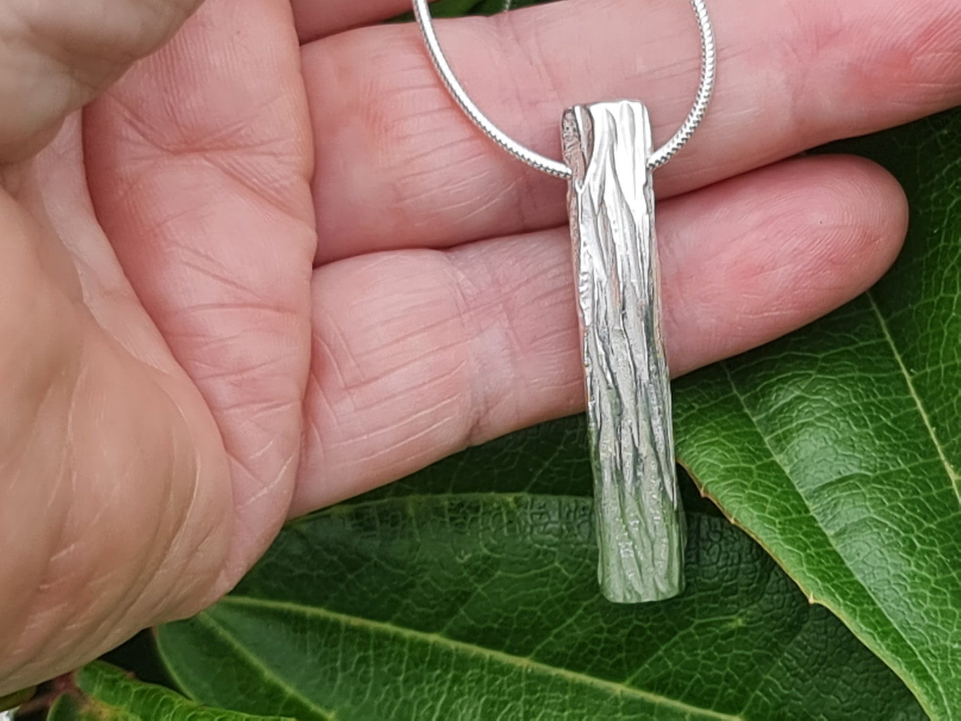 Chunky plus size tree bark textured silver pendant held in the hand against a plant by Kara Jewellery by Charlotte