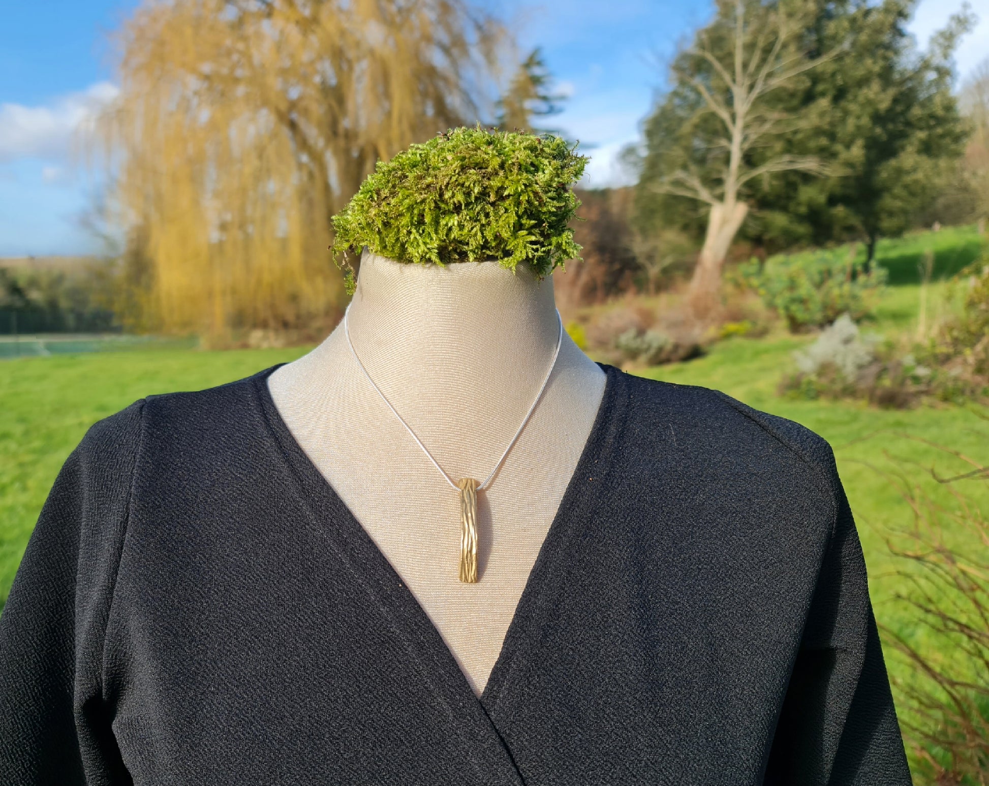 bronze nature inspired curved rectangular pendant on a silver chain.  Shown on a mannequin with a black dress with a garden background