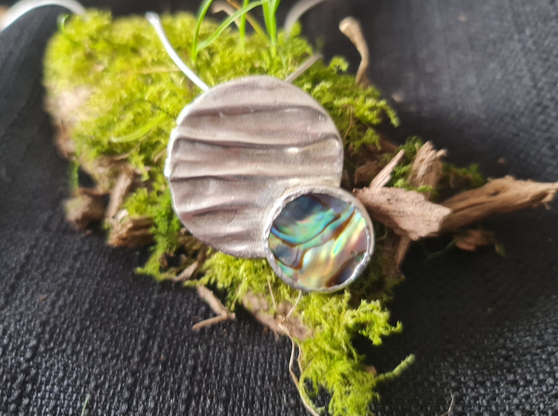 handmade sterling silver rippled pendant with a beautiful blue and green paua shell placed off centre.  The pendant is sitting on a moss cushion.