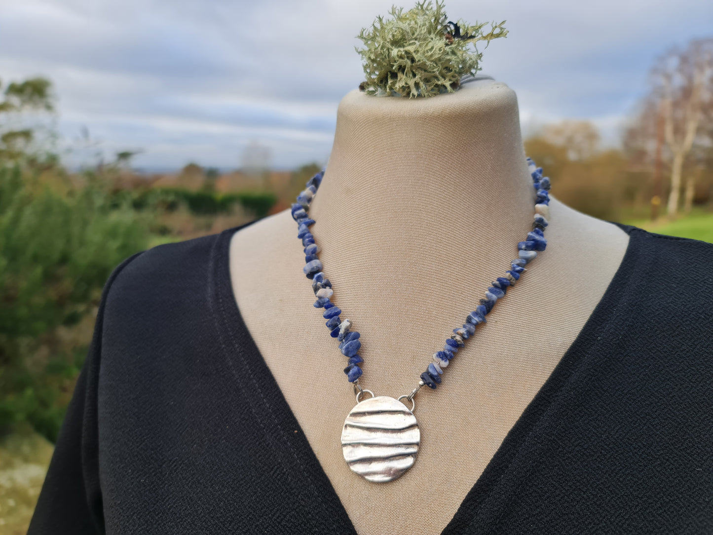 Handmade solid silver 3 mm disc with a ripple texture and blue sodalite beads round the neck.  The pendant is shown on a mannequin wearing a black dress with a country garden background.